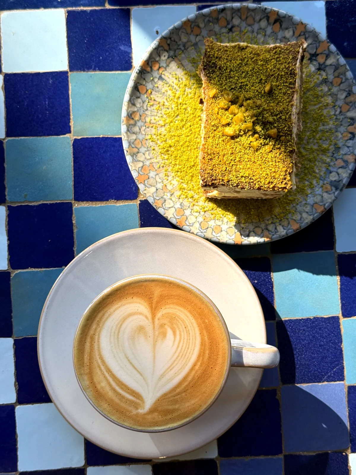 Cappuccino mit Milchschaum-Herz und ein Stück Pistazien-Tiramisu auf einem blauen Mosaiktisch